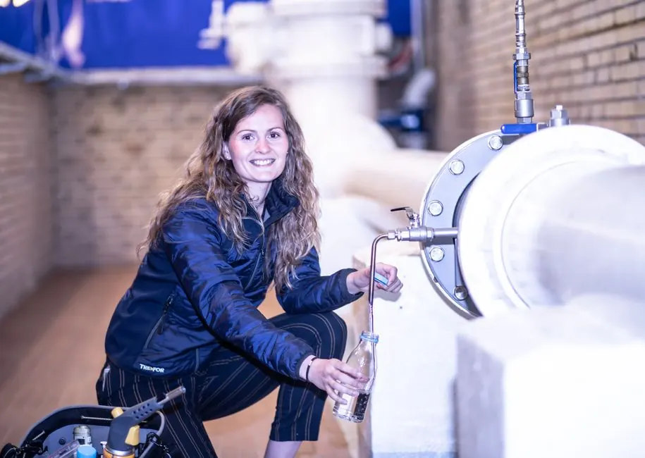 Smiling woman fills a bottle from an industrial pipe in a factory setting.